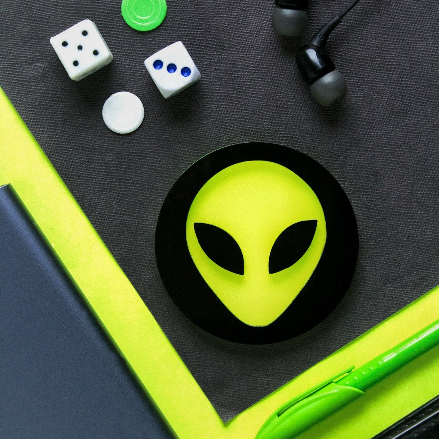 Green alien head sticker on a black surface with dice, earbuds, and a pen.