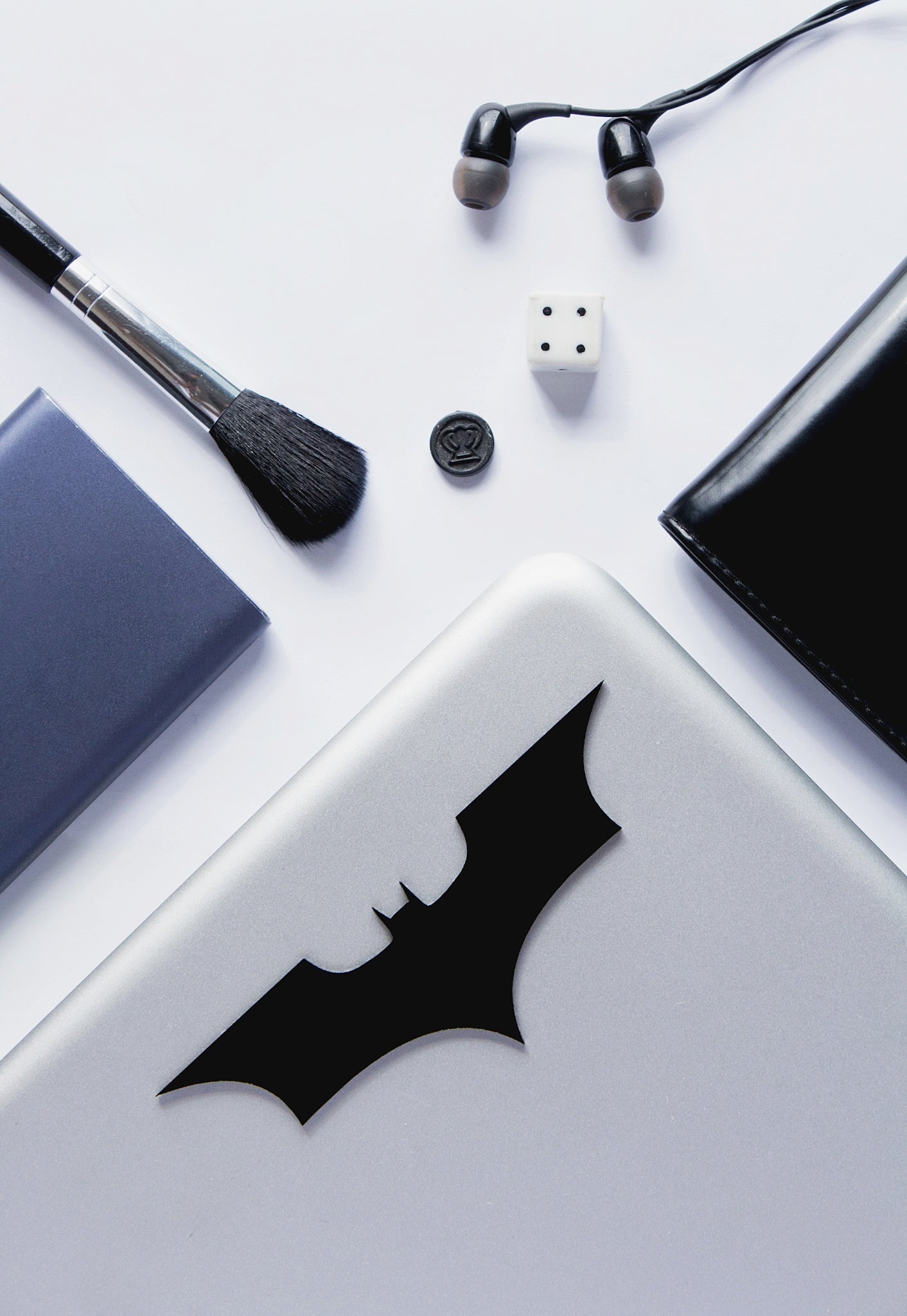 Laptop with Batman logo, makeup brush, earbuds, and dice on a white surface