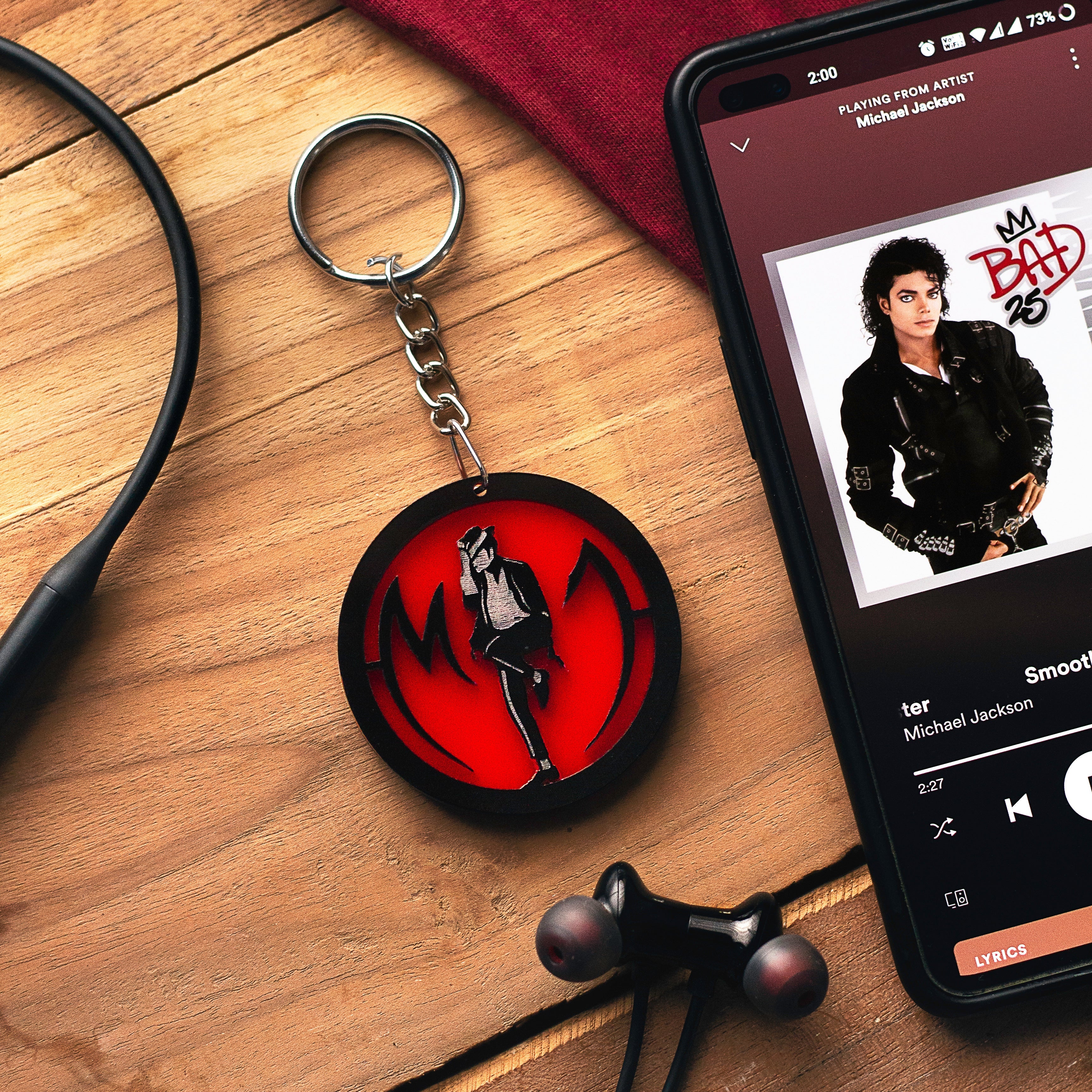 Keychain with a red and black design on a wooden surface next to a phone playing music by Michael Jackson.
