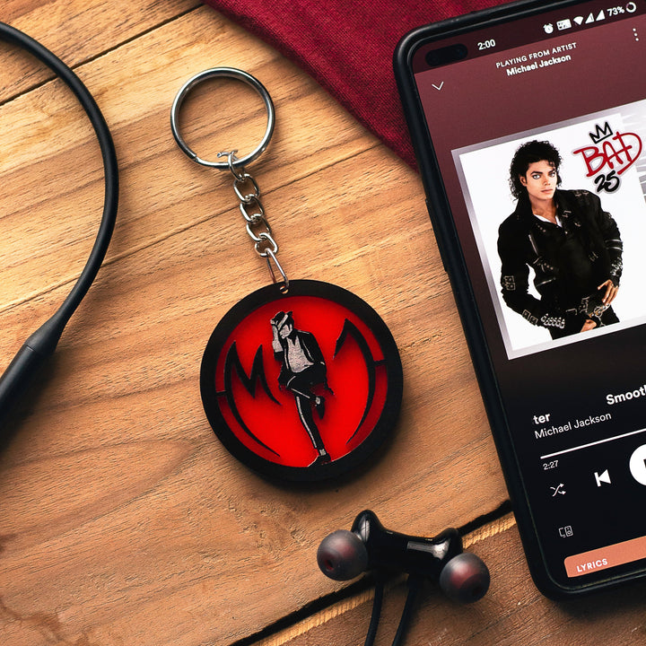 Keychain with a red and black design on a wooden surface next to a phone playing music by Michael Jackson.