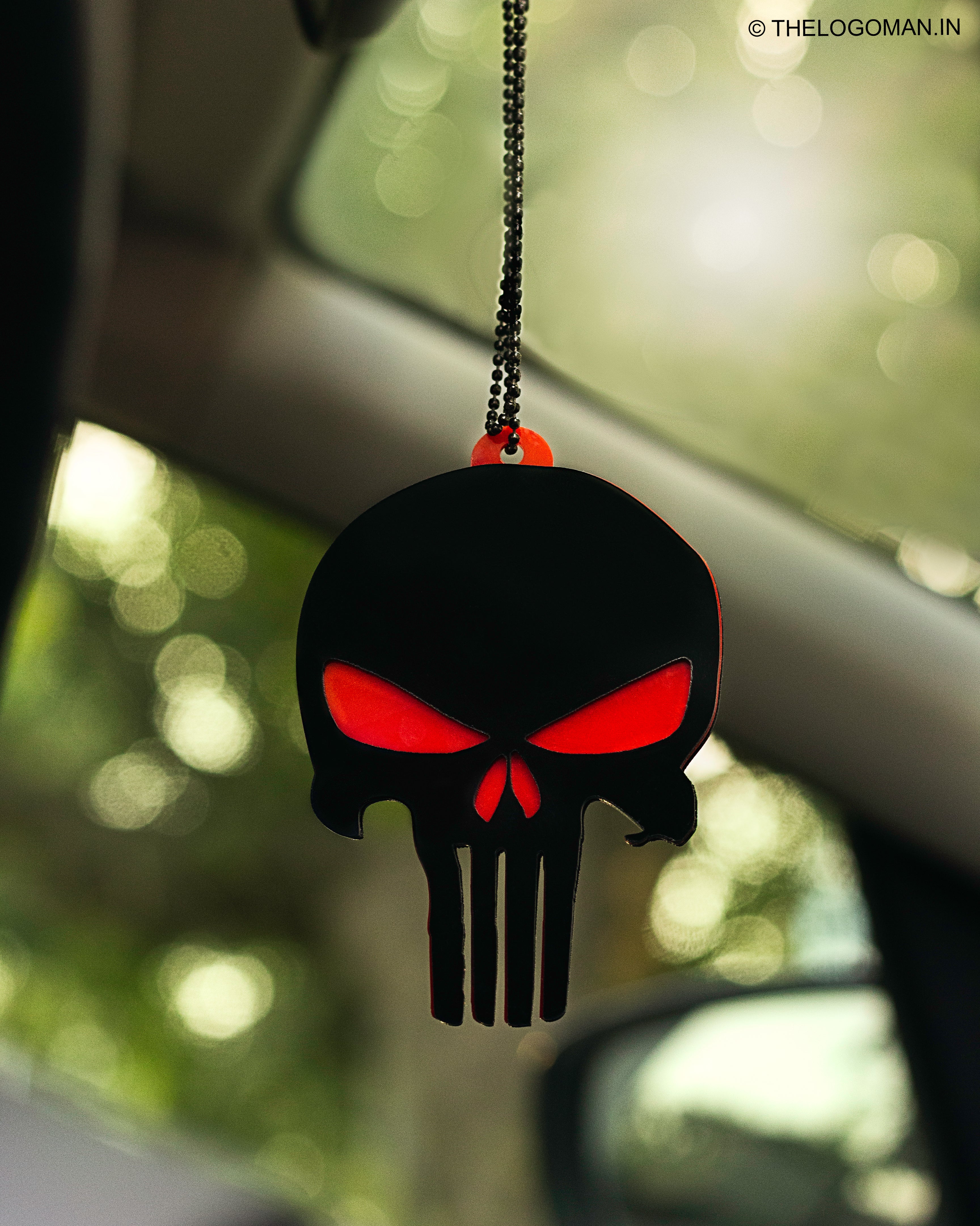 Black and Red Punisher logo car hanging on a car's rearview mirror with blurred greenery in the background.