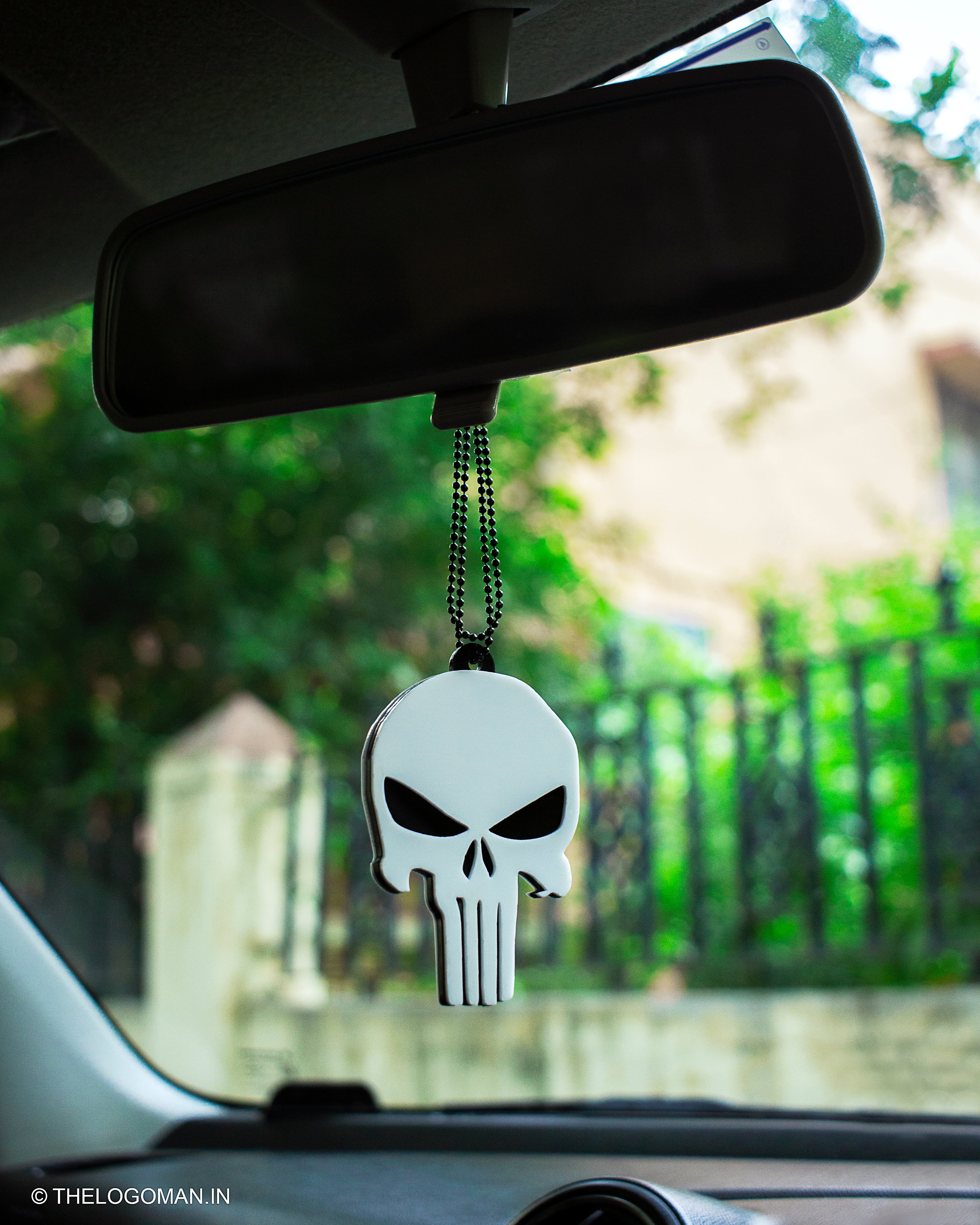 Skull-shaped car hanging from a car's rearview mirror with a blurred outdoor background.